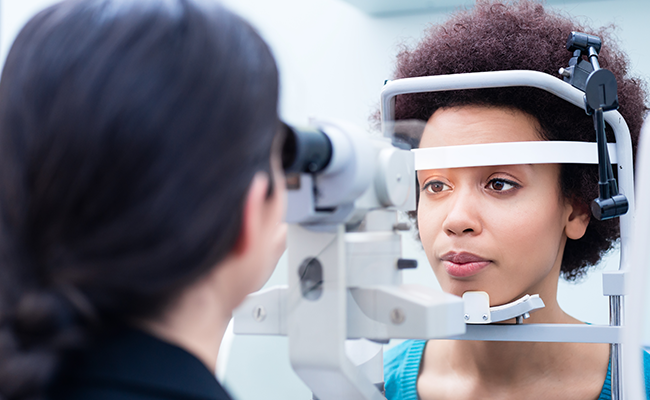 An eye examination in progress with a patient seated at an optical machine while a professional looks into their eyes through a magnifying glass.