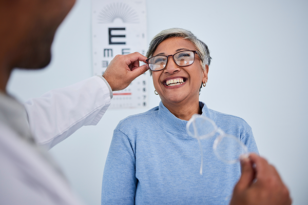 The image shows an elderly woman receiving glasses from an optician, with both individuals smiling and engaged in conversation.
