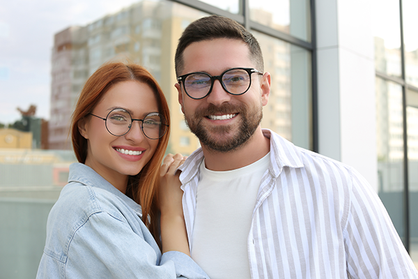 A man and woman posing together, both smiling, with the man wearing glasses and a striped shirt, and the woman with red hair and a white top.